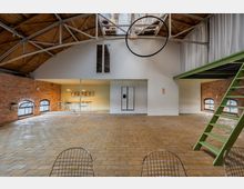 Offener Innenraum eines sanierten Lofts mit Backsteinwänden, großen Rundbogenfenstern und freiliegender Holzbalkendecke. Das Raumdesign umfasst eine Zwischendecke mit grüner Treppe sowie eine offene Küche im Hintergrund.