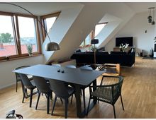 Modernes, helles Wohnzimmer mit Dachschrägen und großen Fenstern, die viel Tageslicht hereinlassen. Im Vordergrund ein Esstisch mit dunkler Platte und sechs Stühlen, dahinter eine schwarze Couch, ein Fernseher und minimalistische Dekoration auf Holzfußboden.