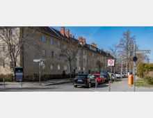 Straßenszene mit einer Reihe von mehrstöckigen Wohngebäuden mit hellbeigen Fassaden und roten Ziegeldächern. Im Vordergrund stehen parkende Autos, Verkehrszeichen wie ein "30-Zone"-Schild sowie ein Gehweg mit Bäumen und einem orangefarbenen Mülleimer.