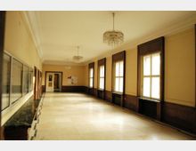 Langer, heller Flur mit cremefarbenen Wänden, großen Fenstern mit dunklen Holzrahmen auf der rechten Seite und einer Fensterbank aus Marmor. Links sind Vitrinen in die Wand eingelassen, und an der Decke hängen mehrere Kronleuchter.