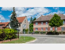 Straßenszene in einem Wohngebiet mit zwei zweistöckigen Häusern, die einfache rote Ziegeldächer und farblich abgesetzte Fassaden besitzen. Im Vordergrund sind ein Gehweg, ein Baum, Straßenschilder sowie Personen mit Kinderwagen zu sehen, umgeben von Gärten mit niedrigen Zäunen.