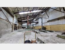 Eine verlassene Industriehalle mit hohen Wänden, einem teils eingestürzten Dach mit sichtbarem Stahlträgerwerk und verschmutztem Boden. An den Wänden sind Rohre, Verkabelungen und gelb gestrichene Bereiche zu sehen, während in der Mitte alte Maschinen und Rohre stehen.