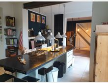 Moderne Wohnküche mit einer großen Kücheninsel aus dunklem Stein, integriertem Herd und Spüle. Im Hintergrund befinden sich Regale an der Wand, eine offene Treppe, Pendelleuchten sowie blaue und weiße Wände mit dekorativen Elementen wie Gemälden und Holzkisten.