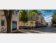 Straßenszene in einem Wohn- und Geschäftsviertel mit gepflasterter Fahrbahn, umgeben von mehrstöckigen Gebäuden in verschiedenen Farben. Im Vordergrund stehen ein Baum und ein Straßenschild, während im Hintergrund Autos und ein Café mit Sitzmöglichkeiten zu sehen sind.