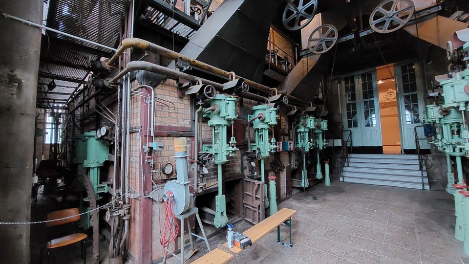 Industriehalle mit historischen Maschinen und Apparaturen, darunter grüne Metallanlagen und verlegte Rohre an einer Ziegelmauer. Im Hintergrund führt eine Treppe zu einer Tür mit gläsernen Fensterpaneelen.