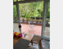 Blick durch eine geöffnete Glastür auf eine Terrasse mit Terrakotta-Boden, auf der mehrere Blumentöpfe mit Pflanzen stehen. Im Hintergrund befinden sich grüne Bäume, und im Vordergrund ist ein Holztisch mit Stuhl, Schreibtischlampe und kleinen Behältern zu sehen.