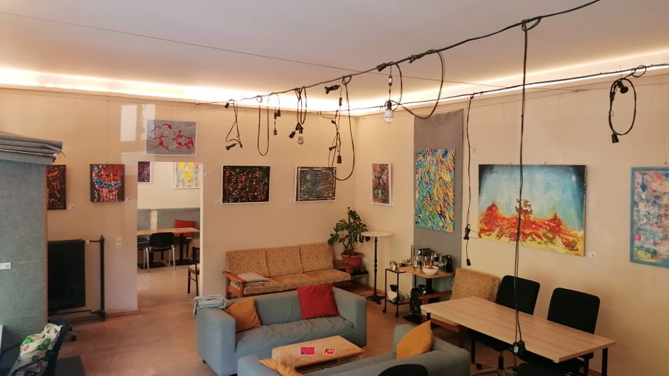 Innenraum eines offenen Atelier- oder Galerieraums mit Sofaecke und mehreren Tischen, an den Wänden hängen viele farbige Gemälde. Von der Decke verlaufen Kabel mit frei hängenden Lampen, im Hintergrund führt ein Durchgang in einen weiteren Raum.