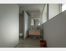 Modernes Badezimmer mit einer schlichten grauen Wandgestaltung, einem wandhängenden Holzwaschtisch, einem großen Spiegel und seitlich einer separaten Toilettenecke. Tageslicht fällt durch ein schmales Fenster an der rechten Wand, ein Holzpapierkorb steht im Vordergrund.