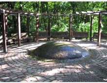 Runder Platz in einem Park mit einem niedrigen, wasserbedeckten Springbrunnen in der Mitte, umgeben von einer kreisförmigen Pflasterfläche, Holzpergolen und Sitzbänken. Im Hintergrund sind Bäume und dichtes Grün zu sehen.