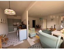 Wohnzimmer mit integrierter Küchenzeile, eingerichtet im Retro-Stil. Die Küche verfügt über grüne Schränke, während das Wohnzimmer helle Sitzmöbel, einen Teppich mit Streifenmuster sowie Dekorationen wie eine Gitarre und kleine Bilderrahmen an der Wand aufweist.