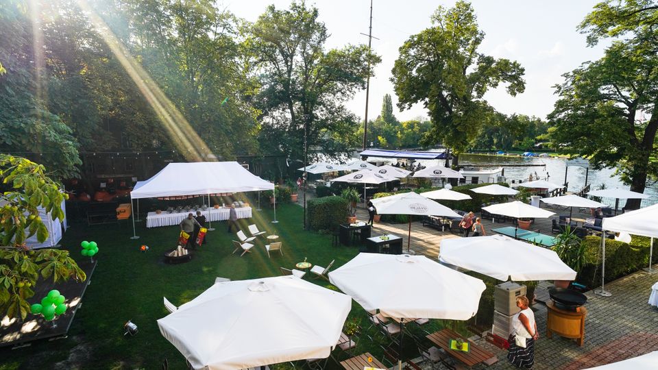 Ein Außenbereich eines Restaurants oder Biergartens an einem Fluss mit weißen Sonnenschirmen, Sitzplätzen aus Holz und einem weißen Zelt für ein Buffet; im Hintergrund sind Bäume und das Wasser sichtbar, mit Stegen und Booten am Ufer.