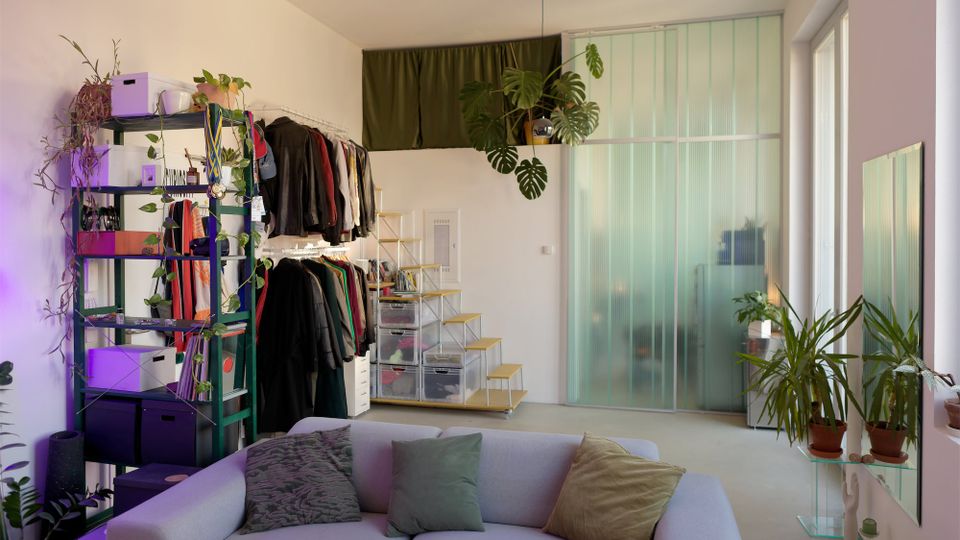 Heller Wohnraum mit Sofa im Vordergrund, offenem Regal und Kleiderstange an der linken Wand sowie einer kleinen Stufenablage mit transparenten Schubladen. Rechts trennt eine hohe, grünlich-transluzente Glaswand den Raum, ergänzt durch große Fenster und mehrere Zimmerpflanzen.