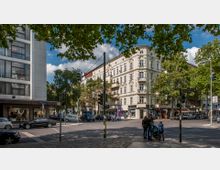 Straßenszene mit einer Kreuzung in einer Stadt, flankiert von mehrstöckigen Gebäuden mit historischen und modernen Fassaden. Im Vordergrund stehen Fußgänger an einer Ampel, umgeben von Laubbäumen, Autos und Geschäften im Erdgeschoss.