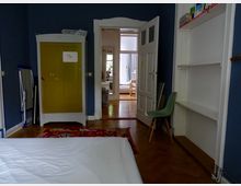 Ein Wohnraum mit dunkelblauen Wänden, einem gelben Kleiderschrank, einer weißen Tür, die in ein helleres Zimmer mit Fenster führt, und Regalen auf der rechten Seite. Im Vordergrund befindet sich ein Bett mit weißer Bettwäsche und ein bunter Teppich auf dem Holzfußboden.