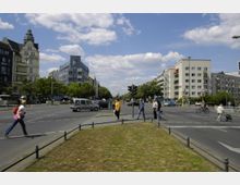 Straßenkreuzung in einer Stadt mit Fußgängern und Autos, flankiert von modernen und historischen Gebäuden. Im Vordergrund eine kleine Grünfläche, im Hintergrund Bäume entlang der Straßen und ein wolkiger Himmel.