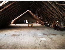 Geräumiger Dachboden mit offenen Holzbalken und Betonboden, teilweise eingerichtet mit gestapelten Stühlen und Tischen. Im Hintergrund ist ein heller Durchgang mit einer Person sichtbar.