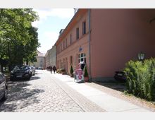 Straßenszene mit Kopfsteinpflaster, linksüberdacht von Bäumen und parkenden Autos. Rechts ist ein langgestrecktes, rosafarbenes Gebäude mit Schaufenstern, vor denen Pflanzentöpfe und ein Werbeschild stehen.