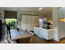 Heller, moderner Küchen- und Essbereich mit großem Holzesstisch, Stühlen und hängenden Glas-Pendelleuchten. Dahinter eine weiße, grifflos gestaltete Einbauküche mit Kücheninsel und dunkler Arbeitsplatte, neben großen Fenstern mit Blick ins Grüne.