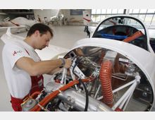 Ein Techniker arbeitet in einer hellen Flugzeugwerkstatt an der Elektronik und den mechanischen Komponenten im Motorbereich eines kleinen Flugzeugs. Im Hintergrund sind weitere Leichtflugzeuge und Werkzeuge zu sehen.