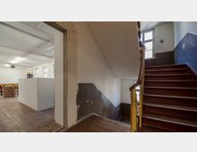 Treppenhaus in einem Altbau mit hölzernen, dunkelroten Stufen und einem gelb gestrichenen Geländer. Links daneben befindet sich ein offener Raum mit Holzdielen, weißen Wänden, einer Sitzgruppe und einer kleinen Theke.