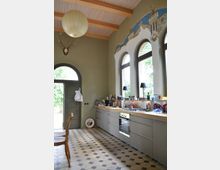 Ein geräumiger Küchenbereich mit hohen Decken und sichtbaren Holzbalken. Die linke Wand enthält eine halbkreisförmige Tür und ein Hirschgeweih, während die rechte Wand durch hohe bogenförmige Fenster mit Glaseinsätzen dominiert wird, über denen ein dekoratives Mosaik mit Türmen und Sternen angebracht ist.