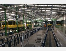 Innenansicht eines Straßenbahn-Depots mit mehreren gelben und blauen Straßenbahnen, umgeben von grünen Metallstützen und offenen Wartungsgruben. Die Halle ist von einem Gitterdach mit viel einfallendem Licht überspannt, und Arbeitsbereiche sind durch Geländer und Plattformen abgegrenzt.