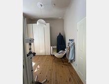 Kinderzimmer mit hellem Holzboden und neutralen Wänden. Es gibt einen weißen Kleiderschrank, ein kleines Babybett mit dunkelblauem Baldachin, einen weißen Schaukelstuhl und hängende Babybekleidung an der Wand.