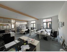 Ein modernes Büro mit Schreibtischen, Stühlen und Computern, viel natürlichem Licht durch große Fenster und sichtbaren Holzbalken an der Decke. An den Wänden hängen gerahmte Bilder, und Regale mit Ordnern und anderen Materialien sind im Hintergrund sichtbar.