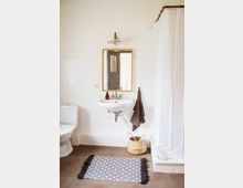 Helles Badezimmer mit weißen Fliesen an der Wand, einem Waschbecken mit Wandspiegel und darüber einer Lampe. Links steht eine Toilette, rechts eine Duschkabine mit weißem Vorhang, ein geflochtener Korb und ein gemusterter Teppich auf dem Boden.