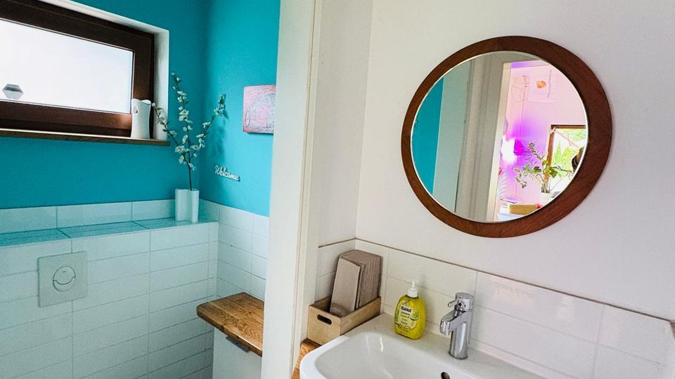 Kleines Badezimmer mit türkisfarben gestrichenen Wänden und weißer Fliesenverkleidung, links mit wandhängendem WC und kleinem Fenster. Rechts ein Waschbecken auf Holzplatte mit rundem Wandspiegel darüber.