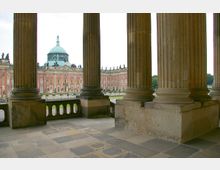 Blick durch eine Reihe von Säulen auf eine barocke Fassade mit einer großen Kuppel. Der Boden besteht aus großen Steinplatten, und im Hintergrund sind Grünflächen und der Himmel sichtbar.