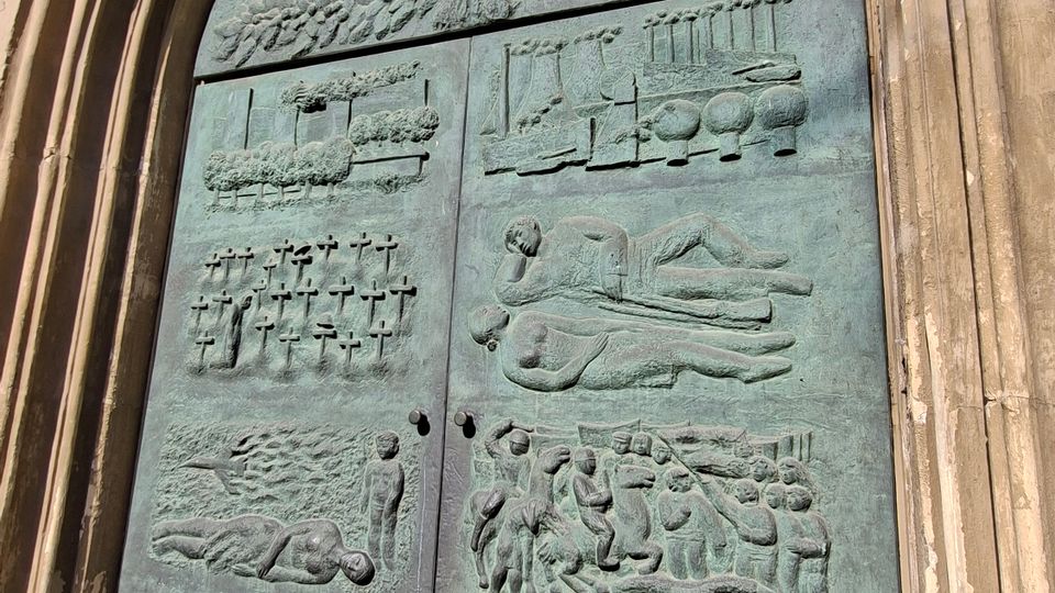 Großes, grün patiniertes Bronzetor mit Reliefdarstellungen, eingefasst von einem hohen, gestuften Spitzbogenportal aus hellem Stein. Das Portal liegt an der Außenfassade eines historischen Gebäudes und steht auf einer flachen Steinstufe mit gepflastertem Boden davor.