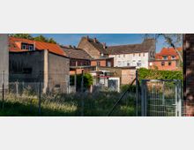 Ein umzäuntes, ungenutztes Grundstück mit bewachsenem Gras im Vordergrund, umgeben von älteren Wohngebäuden mit Ziegeldächern und teils unverputzten Fassaden. Im Hintergrund sind bunte Hauswände und Fenster sichtbar, teilweise mit Balkonen.