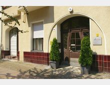 Fassade eines Wohn- oder Bürogebäudes mit gelblichem Putz und roten Fliesen im unteren Bereich. In der Mitte befindet sich eine braune Holztür mit einem bogenförmigen Eingang, flankiert von zwei hohen, grünen Pflanzen in Kübeln; daneben ein Schild mit der Aufschrift „WIRTMANN HAUSVERWALTUNGEN“.