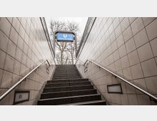 Treppenaufgang einer U-Bahn-Station mit hell gekachelten Wänden und beidseitigen Handläufen, oben ein Schild mit der Aufschrift „U Uhlandstraße“ vor kahlen Baumkronen.