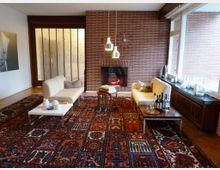 Wohnzimmer mit gemustertem Teppichboden, einem gemauerten Kamin an einer Ziegelwand, zwei cremefarbenen Sofas, und niedrigen Tischen mit Dekorationen. Große Fenster rechts lassen natürliches Licht herein, während links eine Holzschiebetür und ein Kunstwerk sichtbar sind.