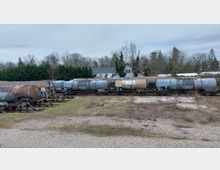 Güterzug mit mehreren Kesselwagen auf einem Gleis in einer ländlichen Umgebung, umgeben von Büschen, Bäumen und Häusern im Hintergrund. Im Vordergrund ist ein mit Kopfsteinpflaster und Gras bewachsener Bereich zu sehen.