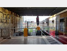 Bahnsteig unter einer Brücke mit einer stehenden S-Bahn auf der rechten Seite. Im Hintergrund sind Bahngleise, ein Signal, Baukräne sowie Graffiti an der Brückenwand zu sehen.