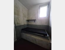 Kleiner, einfacher Raum mit abgenutzten, gestrichenen Wänden in Grau und Weiß. Der Raum enthält eine schmale, leicht erhöhte Liege mit schwarzer Matratze und ein kleines Fenster mit Gitter, das Licht hereinlässt; ein schlichtes Regal hängt an der Wand.