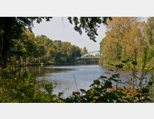 Flusslandschaft mit dichtem Baumbestand an beiden Ufern und einer grünen Bogenbrücke im Hintergrund. Im Vordergrund sind Büsche und Zweige sichtbar, die teilweise die Sicht auf das Wasser einrahmen.