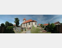 Fachwerkkirche mit roten Dachziegeln in einer ländlichen Umgebung, umgeben von einem Friedhof mit Grabsteinen und gepflegten Gräbern. Im Hintergrund sind Bäume, vereinzelt Gebäude und eine Ziegelmauer sichtbar.