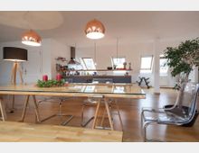 Helles Wohnzimmer mit moderner, offener Küchenzeile, großen Dachfenstern und einem langen Holztisch mit Glasplatte, umgeben von transparenten Kunststoffstühlen. Im Raum befinden sich Kupfer-Pendelleuchten, eine Stehlampe und eine große grüne Zimmerpflanze.