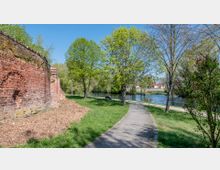 Eine gepflasterte Gehwegführung durch eine Grünanlage mit Bäumen und Sträuchern, links eine rote Backsteinmauer, rechts ein Fluss mit Geländer und eine Sitzbank im Hintergrund. Der Himmel ist klar und blau.