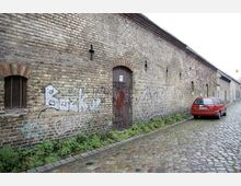 Eine lange Backsteinmauer mit Graffiti übersät, entlang einer gepflasterten Straße. An der Mauer befinden sich eine Holztür und ein kleines, vergittertes Fenster; ein rotes Auto ist am rechten Straßenrand geparkt.