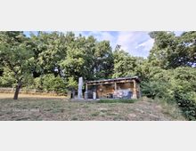 Kleines, modernes Holzhaus mit flachem Vordach und großer Glasfront auf einer Lichtung, davor eine Terrasse mit Außenmöbeln. Umgeben von dichtem Wald und Sträuchern, mit leicht abfallender Wiese im Vordergrund.