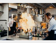 Eine professionelle Restaurantküche mit zwei Köchen in weißen Kochjacken und schwarzen Schürzen. Einer der Köche steht neben einem Herd, auf dem eine große Flamme aufsteigt; Küchenutensilien hängen von einer Deckenhalterung, und die Arbeitsfläche ist mit Gewürzflaschen und Kochwerkzeugen bestückt.