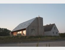 Modernes Wohnhaus mit einer Holzfassade und großen Glasfenstern, kombiniert mit einem angrenzenden älteren Gebäude mit grauer Putzfassade und Satteldach. Im Vordergrund ist eine Straße, umgeben von Wildwiese und vereinzelten Bäumen im Hintergrund.