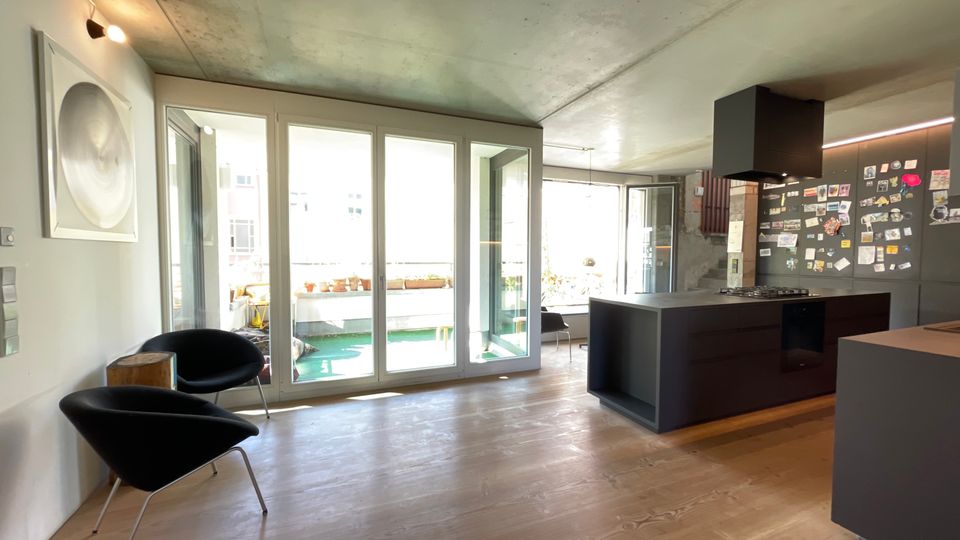 Moderner Wohnraum mit offener Küche, großen Fenstern und Blick auf einen Balkon mit Pflanzen. Die Küche ist in dunklen Farben gehalten, mit einer zentralen Insel, während der Boden aus hellem Holz besteht.