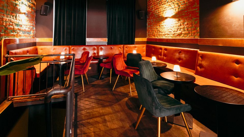 Gemütlicher Lounge- und Barbereich mit freigelegten Ziegelwänden, gepolsterten roten Sitzbänken entlang der Wände und kleinen runden Tischen mit Tischlampen. Samtstühle in Rot und Dunkelgrün stehen auf dunklem Boden, im Hintergrund hängen schwarze Vorhänge und an den Wänden sind Leuchten und Lautsprecher montiert.