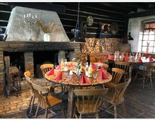 Gemütlicher Gastraum in rustikalem Stil mit dunklen Holzbalken, einem großen gemauerten Kamin, Holzstapeln an der Wand und Holztischen, die mit roten Stoffservietten und Getränken gedeckt sind. Der Raum ist mit hölzernen Stühlen und traditioneller Dekoration ausgestattet.